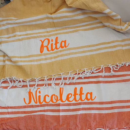 Una pila di asciugamani colorati In Casa di Ale Telo Mare - FOUTA è disposta sulla sabbia vicino a una stella marina e a sassi levigati, con il mare sullo sfondo. Ogni asciugamano è etichettato in italiano da Arancione a Fucsia.