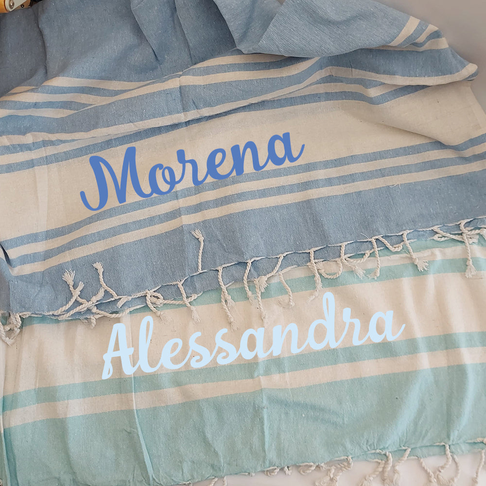 Una pila di asciugamani colorati In Casa di Ale Telo Mare - FOUTA è disposta sulla sabbia vicino a una stella marina e a sassi levigati, con il mare sullo sfondo. Ogni asciugamano è etichettato in italiano da Arancione a Fucsia.