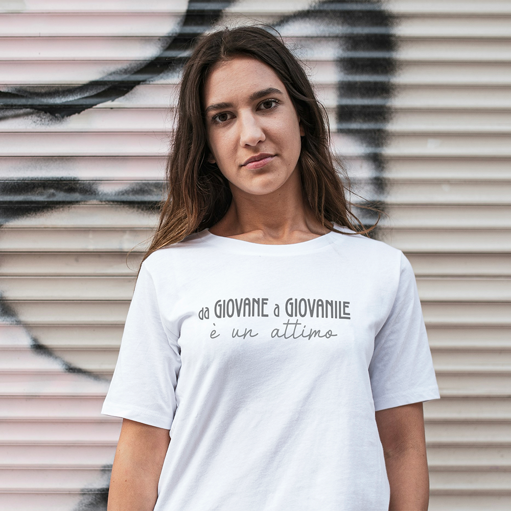Una donna indossa la T-shirt Giovane by Il mio negozio, con testo in italiano, mentre si trova di fronte a un murale con colori pastello e linee nere. t-shirt personalizzata