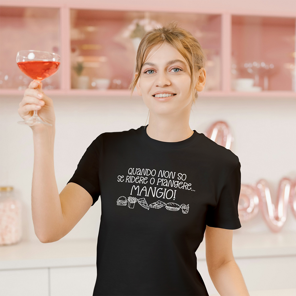 La T-shirt Mangio de Il mio negozio è caratterizzata dal testo in italiano "Quando non so se ridere o piangere... MANGIO!" e simpatica food art. Maglietta nera divertente, ideale come regalo per chi ama le magliette personalizzate. Mostrato su superficie in legno chiaro.