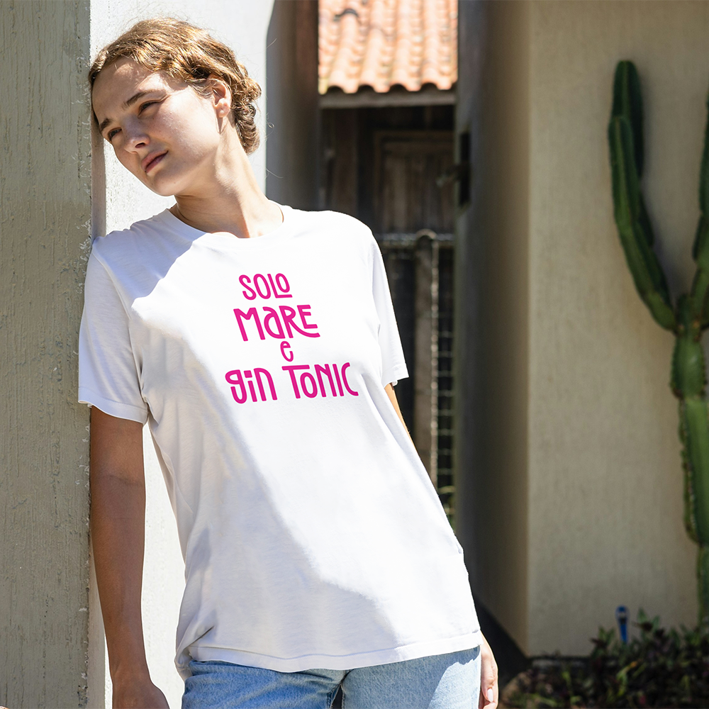 La T-shirt Mare e GinTonic de Il mio negozio, bianca con scritta rosa "SOLO MARE & GIN TONIC", è caratterizzata da maniche arrotolate e orlo annodato, perfetta per chi ama le magliette personalizzate divertenti. Accanto a un cappello intrecciato su sfondo chiaro.