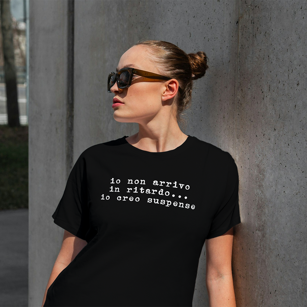 Una donna con gli occhiali da sole si appoggia a un muro indossando la T-shirt Ritardo di Il mio negozio, con la scritta "Io non arrivo in ritardo... io creo suspense". Un'idea regalo unica per chi ama distinguersi.
