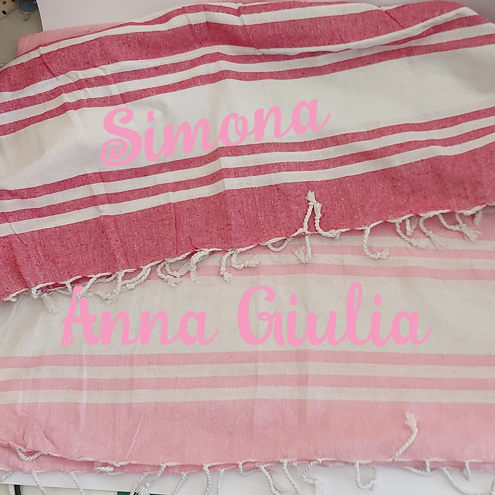 Una pila di asciugamani colorati In Casa di Ale Telo Mare - FOUTA è disposta sulla sabbia vicino a una stella marina e a sassi levigati, con il mare sullo sfondo. Ogni asciugamano è etichettato in italiano da Arancione a Fucsia.