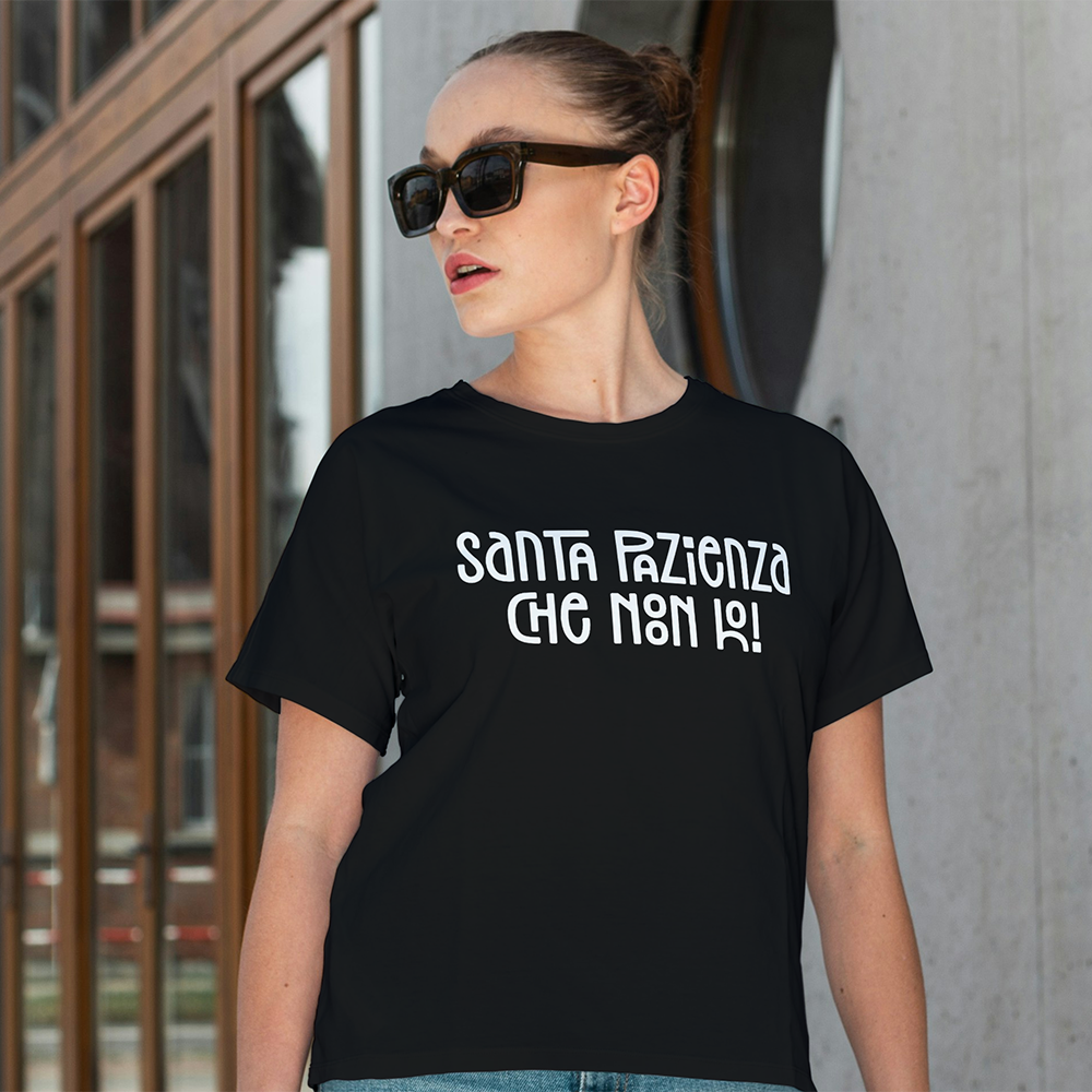 Una donna con gli occhiali da sole indossa la "T-shirt Santa" de Il mio negozio, con la frase "Santa Pazienza che non ho!", mentre si trova all'aperto.