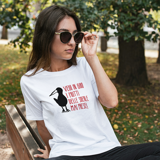 Una donna con occhiali da sole e una T-shirt Sberle by Il mio negozio, con un uccello e un testo in italiano, siede su una panchina del parco.