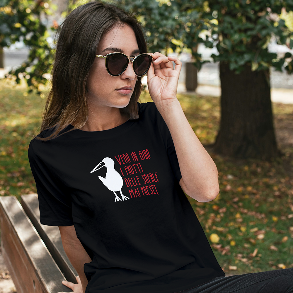Una donna con occhiali da sole e una T-shirt Sberle by Il mio negozio, con un uccello e un testo in italiano, si siede su una panchina all'aperto.