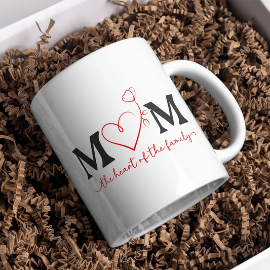 Tazza bianca dedicata alla mamma con scritta Mom The Heart of the Family, appoggiata in confezione regalo con carta kraft.