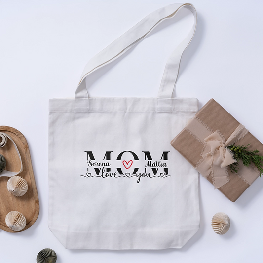 Tote bag bianca personalizzata per la mamma con nomi e dedica, fotografata accanto a confezione regalo.