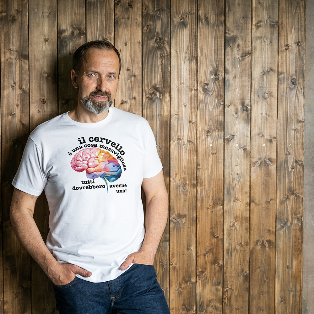 La T-shirt bianca Cervello de Il mio negozio, con un cervello colorato e la frase "Il cervello è una cosa meravigliosa, tutti dovrebbero averne uno!", è appoggiata vicino agli occhiali da sole su una superficie strutturata: una divertente maglietta personalizzata!.