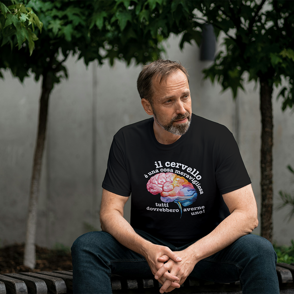 La T-shirt bianca Cervello de Il mio negozio, con un cervello colorato e la frase "Il cervello è una cosa meravigliosa, tutti dovrebbero averne uno!", è appoggiata vicino agli occhiali da sole su una superficie strutturata: una divertente maglietta personalizzata!.