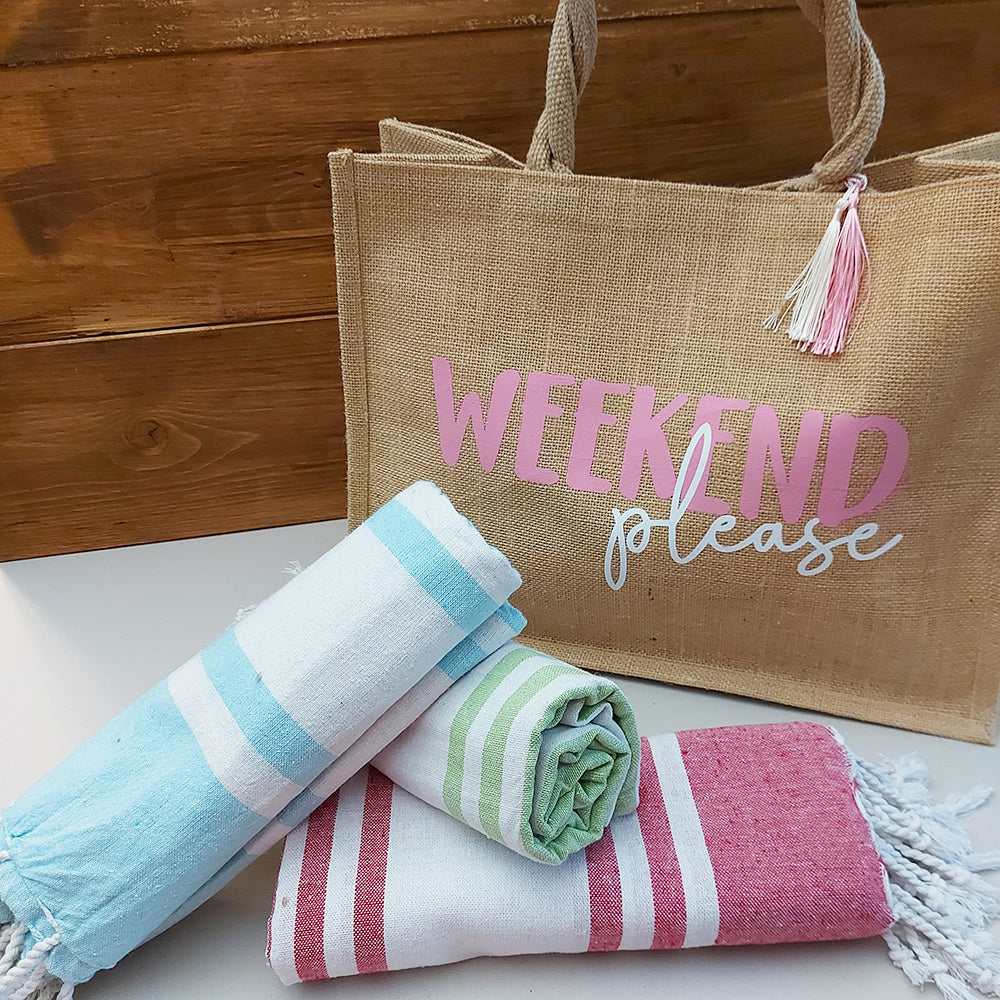 Il Combo Borsa Juta e Telo Fouta WEEKEND di In Casa di Ale è composto da una borsa in juta color tan con la scritta "WEEKEND please" in rosa/bianco e da un asciugamano fouta a righe rosse, ideale per le fughe estive del 2025.