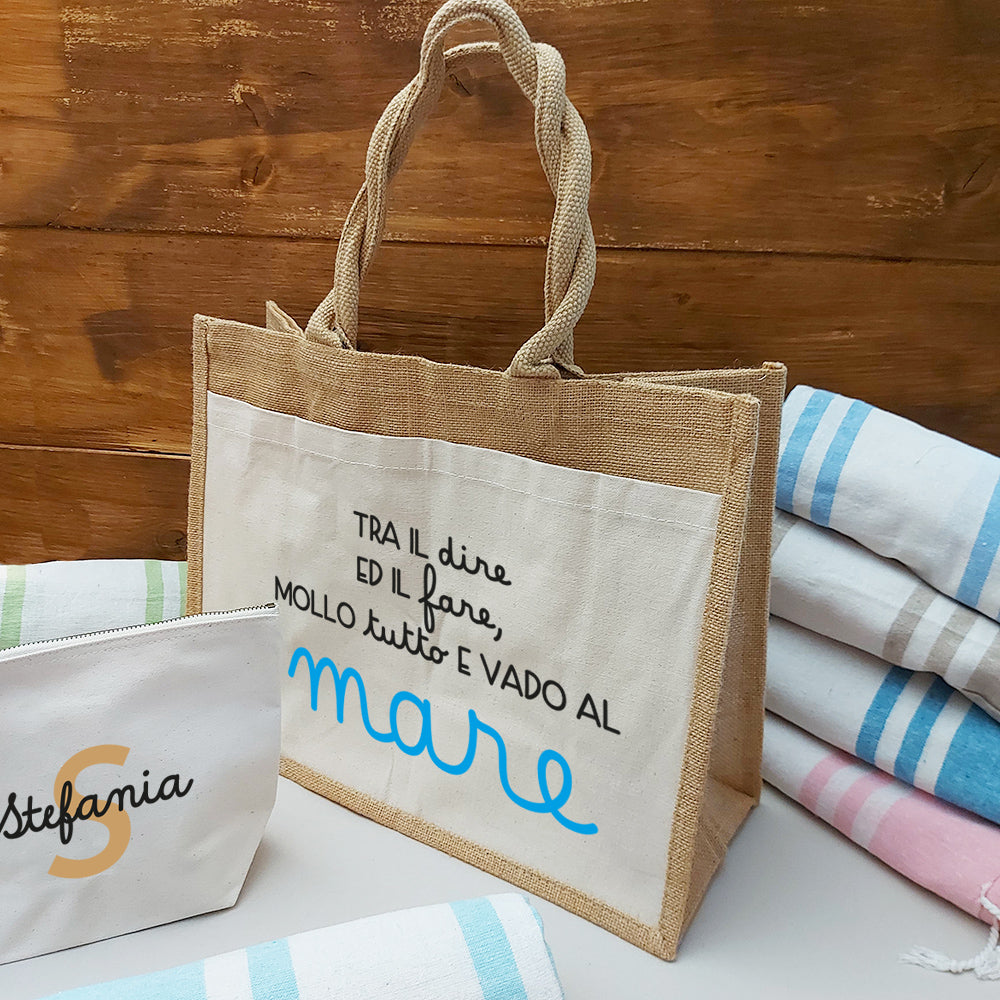 Il Combo Borsa Juta, Telo Fouta e Pochette di Casa di Ale presenta una borsa in juta con testo in italiano e scritte in blu, un sacchetto con zip Stefania bianco con una S arancione e un Telo Fouta a righe, il tutto su una superficie chiara con uno sfondo in legno.