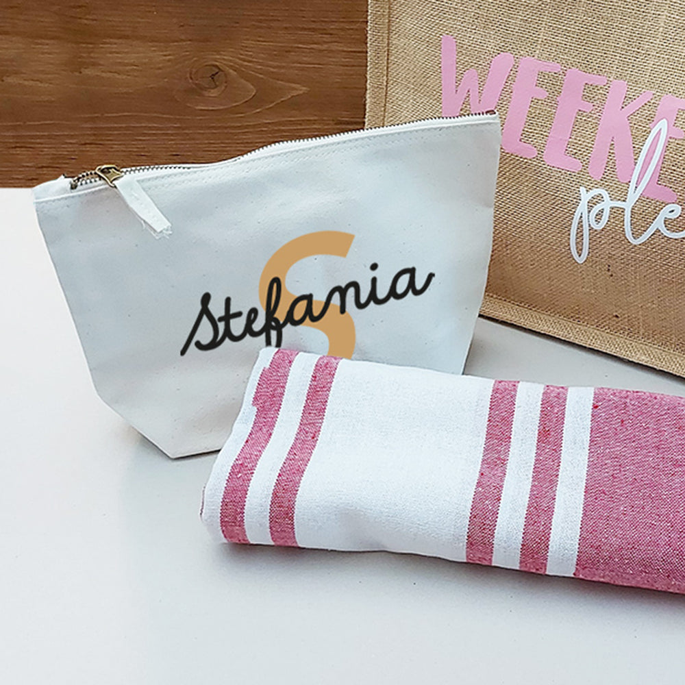 Il Combo Borsa Juta, Telo Fouta e Pochette di Casa di Ale presenta una borsa in juta con testo in italiano e scritte in blu, un sacchetto con zip Stefania bianco con una S arancione e un Telo Fouta a righe, il tutto su una superficie chiara con uno sfondo in legno.