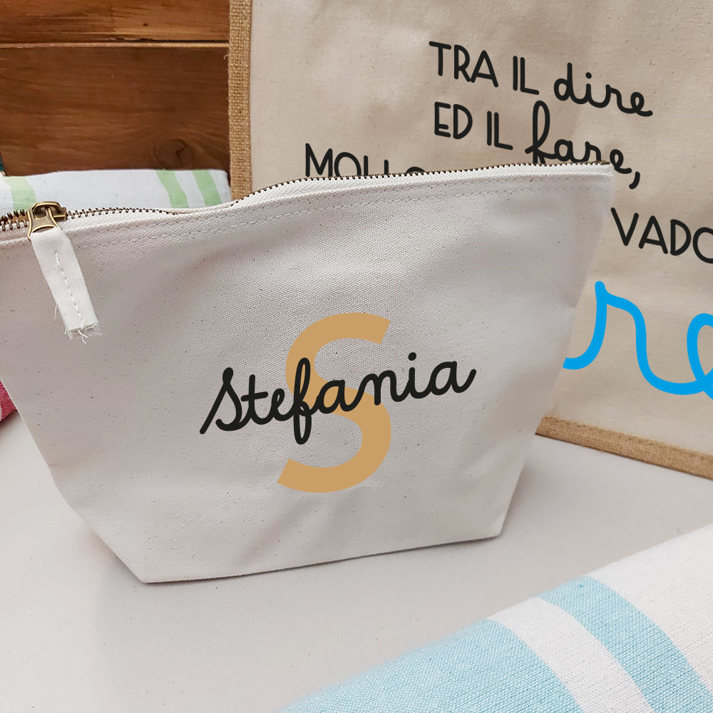 Il Combo Borsa Juta, Telo Fouta e Pochette di Casa di Ale presenta una borsa in juta con testo in italiano e scritte in blu, un sacchetto con zip Stefania bianco con una S arancione e un Telo Fouta a righe, il tutto su una superficie chiara con uno sfondo in legno.