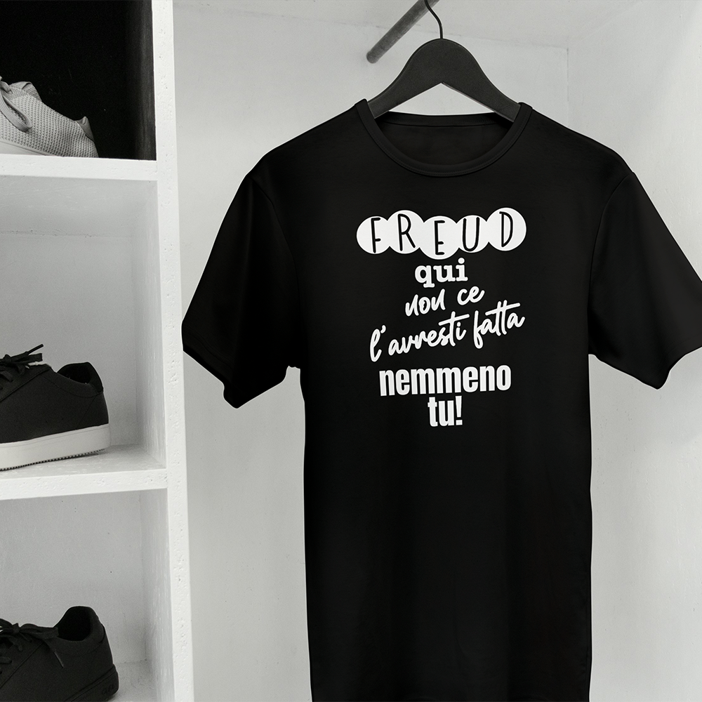 La T-shirt Freud de Il mio negozio è una maglietta bianca ripiegata con il testo italiano "Freud qui non ce l'hai fatta nemmeno tu!" visualizzato su una superficie nera; sullo sfondo compare una tazza rosa con puntini neri, che la rende una perfetta idea regalo.