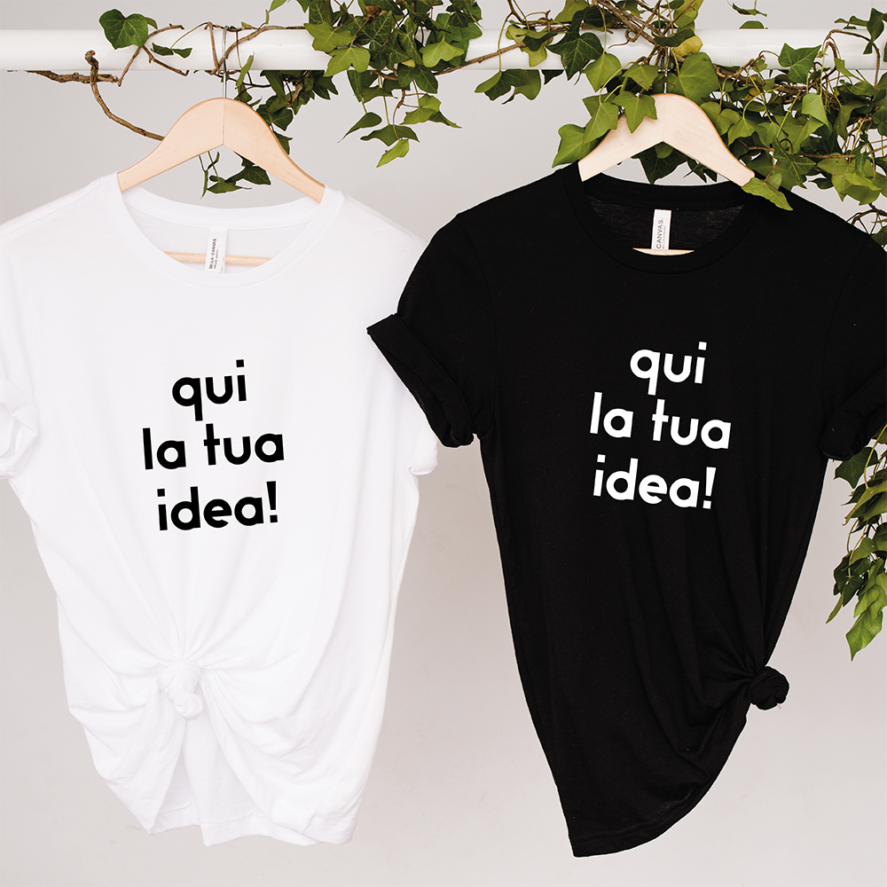 Due "T-shirt da personalizzare" de Il mio negozio sono appese a grucce di legno con sopra un'edera verde. Le magliette bianche e nere mostrano "qui la tua idea!"-perfette come magliette personalizzate divertenti o idee regalo originali.