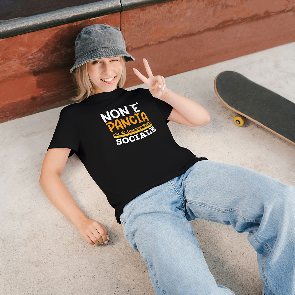 Una persona sorridente in jeans e cappello a secchiello si trova accanto a uno skateboard, facendo il segno della pace e mostrando la nostra T-shirt Pancia by Il mio negozio, ideale per chi cerca un regalo originale.