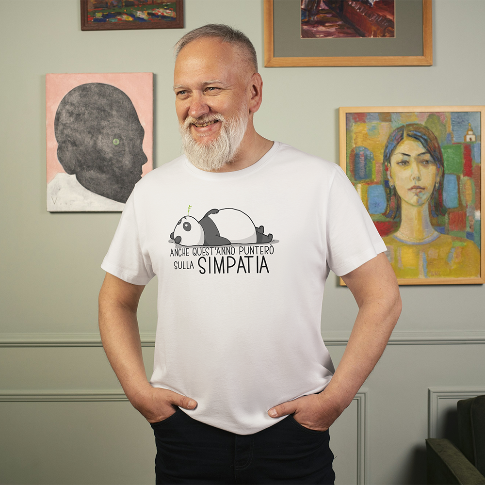 Un uomo sorridente con la barba bianca indossa la T-shirt Simpatia de Il mio negozio, con un panda a fumetti e una scritta in italiano, accanto a un'opera d'arte colorata.