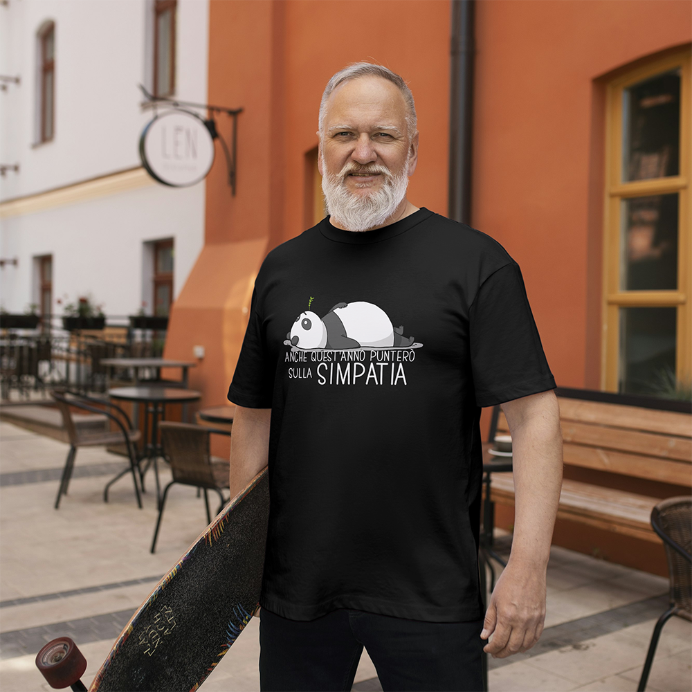 Un uomo anziano sorridente con la barba grigia tiene in mano uno skateboard e indossa la T-shirt Simpatia de Il mio negozio, con il disegno di un panda, all'aperto vicino a un caffè.
