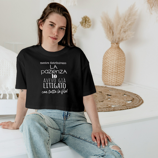 Una donna che indossa la T-shirt Pazienza di Il mio negozio, con testo in italiano, siede su un letto in una stanza luminosa e accogliente dall'arredamento neutro.