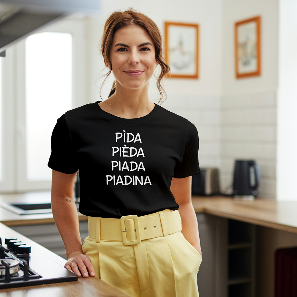 Una donna sorridente in cucina indossa la T-shirt Piadina de Il mio negozio, con la scritta "PIDA, PIEDA, PIADA, PIADINA", a dimostrazione di come le magliette personalizzabili siano un regalo divertente e originale.