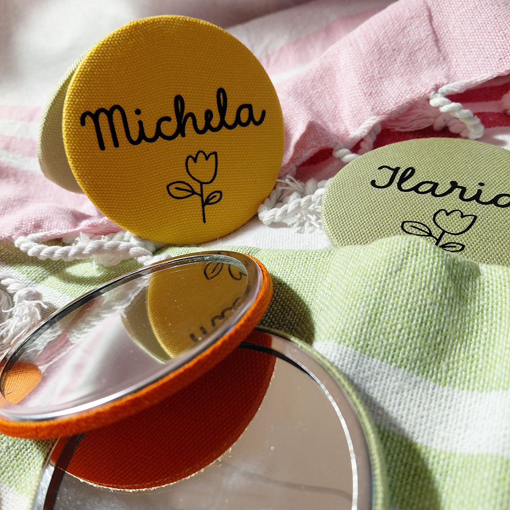 Due badge rotondi in tessuto, uno raffigurante Michela con un tulipano, e gli specchietti tondi colorati e personalizzati di In Casa di Ale, disegnati con tessuti a righe e rosa.