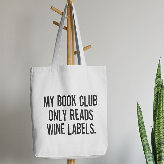 La Tote bag con illustrazioni di Il mio negozio, stampata con "MY BOOK CLUB ONLY READS WINE LABELS", appesa a un attaccapanni, è il regalo originale in cotone perfetto per chi ama libri e vino.