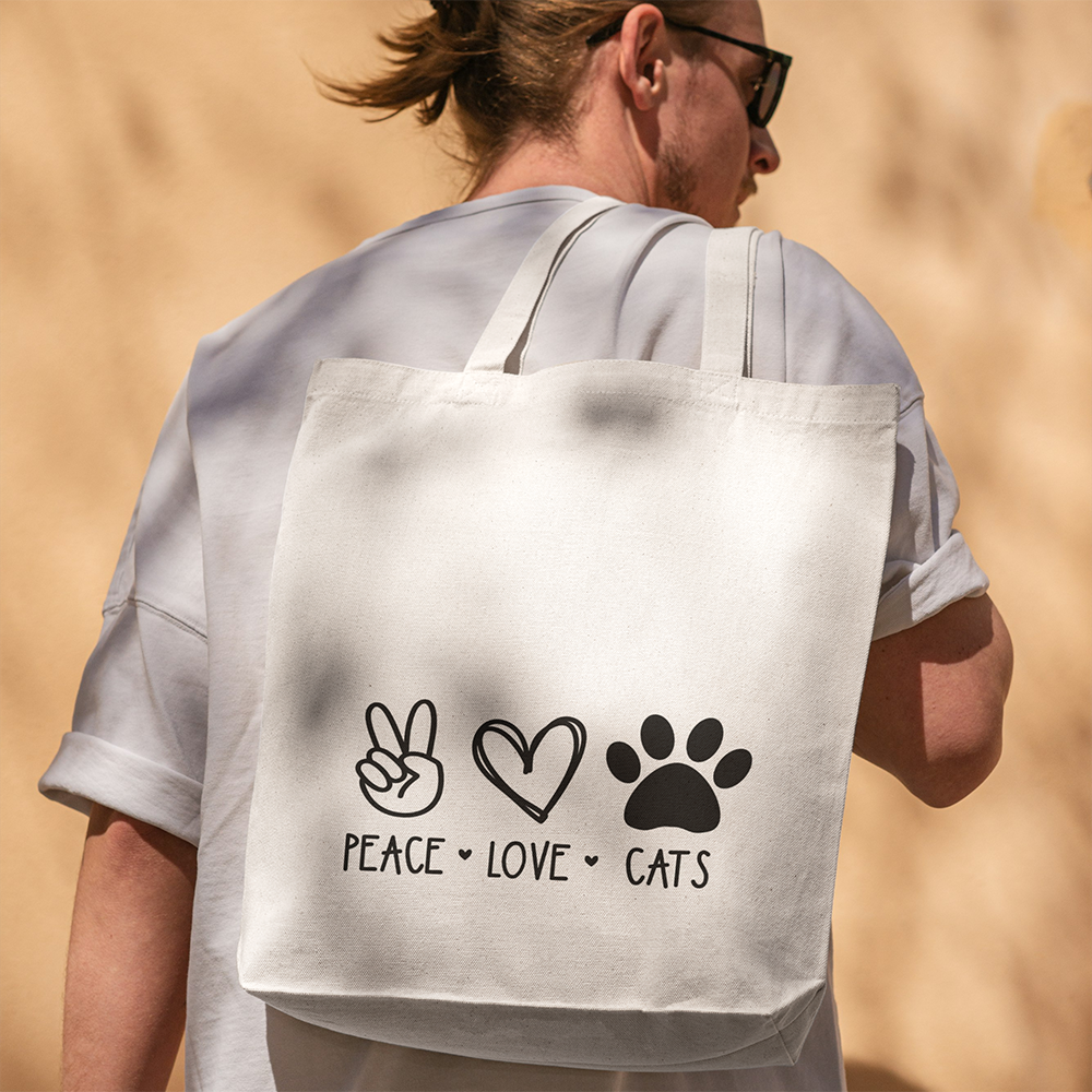 Una persona con occhiali da sole e camicia bianca porta con sé la borsa Tote con illustrazioni de Il mio negozio, con icone nere della pace, del cuore e della zampa con la scritta "Peace Love Cats" - un regalo originale e personalizzato per gli amanti dei gatti.