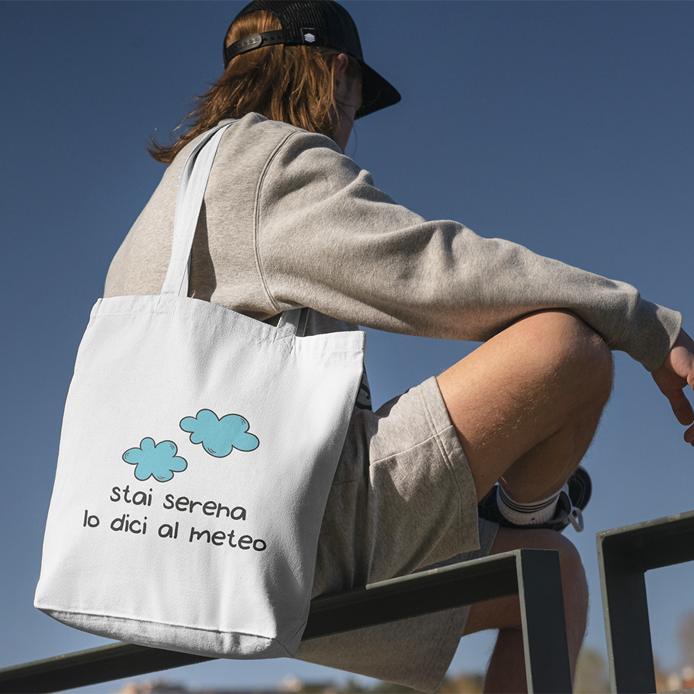 Una persona in abiti casual e berretto siede all'aperto su una ringhiera di metallo, portando con sé la Tote bag con illustrazioni de Il mio negozio, decorata con due nuvole blu e la frase italiana "stai serena lo dici al meteo".