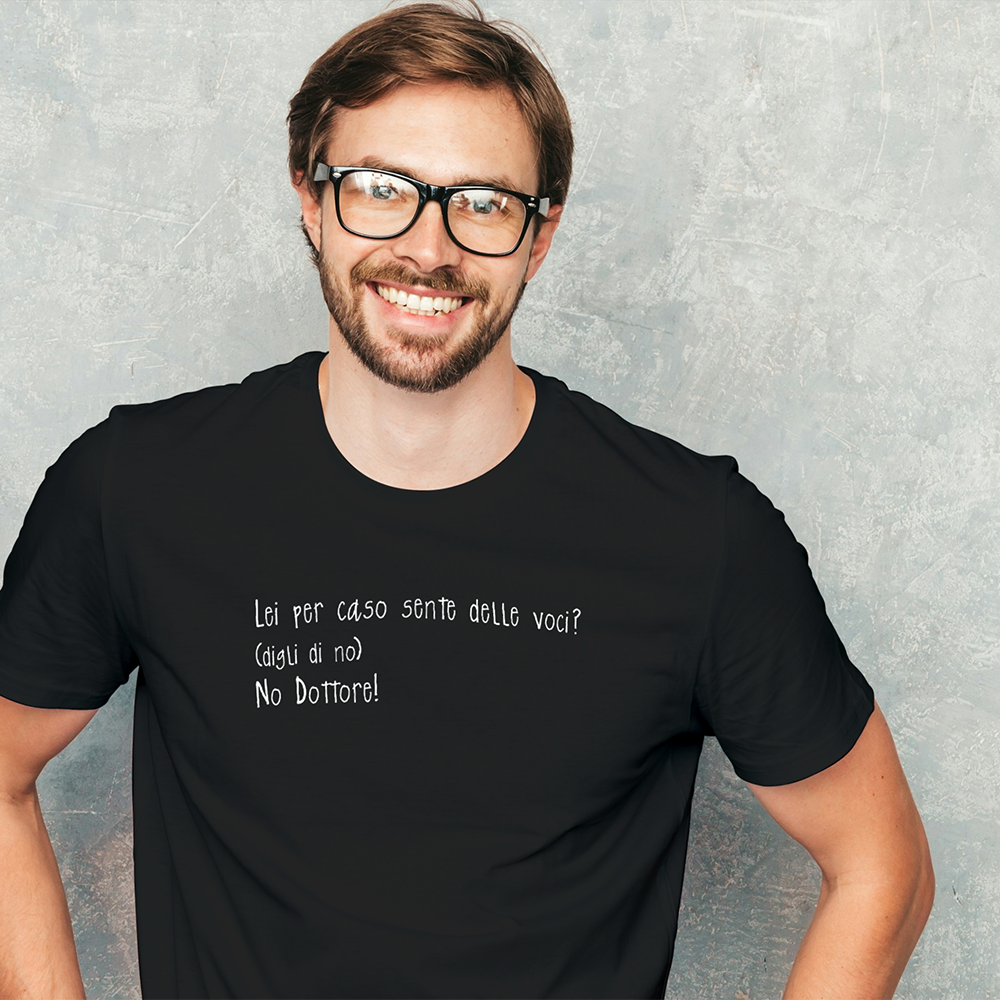 Uomo sorridente con occhiali che indossa una maglietta Il mio negozio "Sento voci" con testo italiano, in piedi contro un muro grigio chiaro.