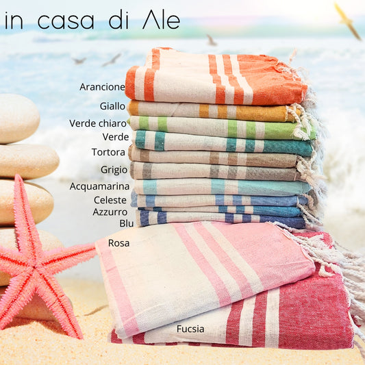 Una pila di asciugamani colorati In Casa di Ale Telo Mare - FOUTA è disposta sulla sabbia vicino a una stella marina e a sassi levigati, con il mare sullo sfondo. Ogni asciugamano è etichettato in italiano da Arancione a Fucsia.