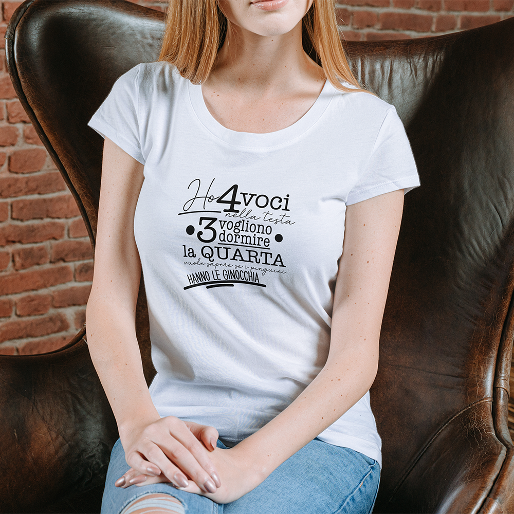La T-shirt 4 voci de Il mio negozio è appesa a una staccionata di legno. Questa maglietta bianca personalizzata riporta in nero il testo italiano: Ho 4 voci nella testa. 3 vogliono dormire, la quarta vuole sapere se i pinguini hanno le ginocchia.