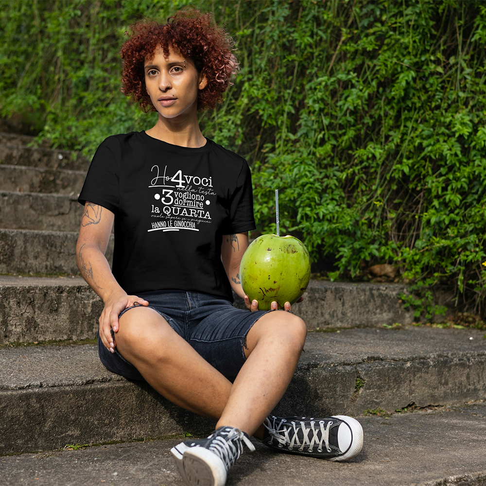 La T-shirt 4 voci de Il mio negozio è appesa a una staccionata di legno. Questa maglietta bianca personalizzata riporta in nero il testo italiano: Ho 4 voci nella testa. 3 vogliono dormire, la quarta vuole sapere se i pinguini hanno le ginocchia.