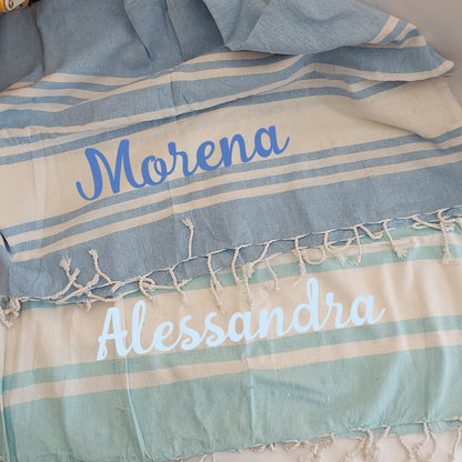 Una pila di asciugamani colorati In Casa di Ale Telo Mare - FOUTA è disposta sulla sabbia vicino a una stella marina e a sassi levigati, con il mare sullo sfondo. Ogni asciugamano è etichettato in italiano da Arancione a Fucsia.