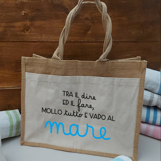 Borsa in juta con grande tasca frontale in canvas e scritta Tra il dire ed il fare, mollo tutto e vado al mare, abbinata a teli fouta colorati.