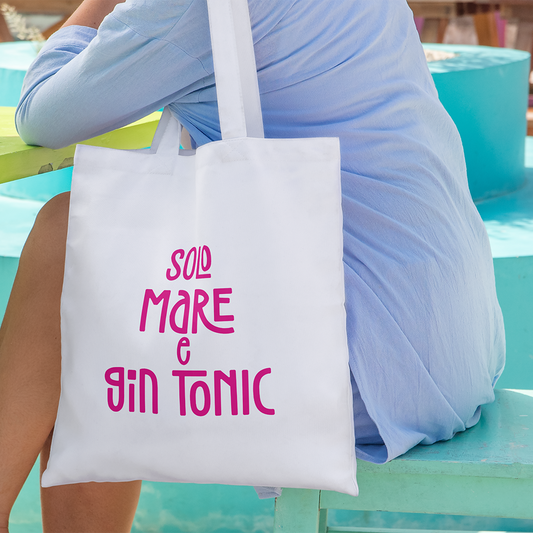 Una donna in abito azzurro siede con la tote bag personalizzata de Il mio negozio, con la scritta "Solo mare e gin tonic" in rosa, che evidenzia un design unico.