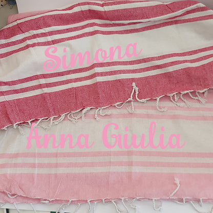 Una pila di asciugamani colorati In Casa di Ale Telo Mare - FOUTA è disposta sulla sabbia vicino a una stella marina e a sassi levigati, con il mare sullo sfondo. Ogni asciugamano è etichettato in italiano da Arancione a Fucsia.