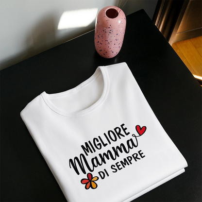 T-shirt bianca per la mamma con scritta Migliore Mamma di sempre, piegata accanto a vaso rosa su tavolo scuro.