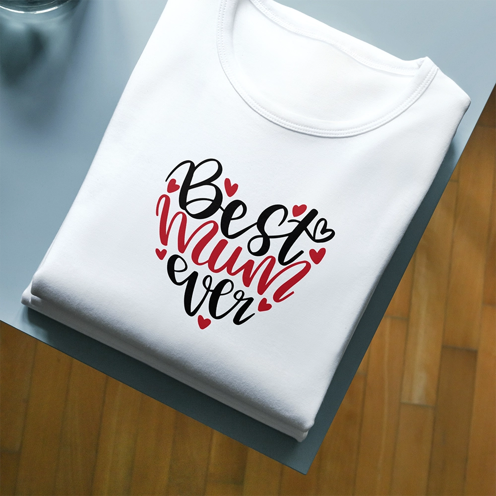 T-shirt bianca per la mamma con scritta Best Mum Ever e piccoli cuori rossi, piegata su piano scuro.
