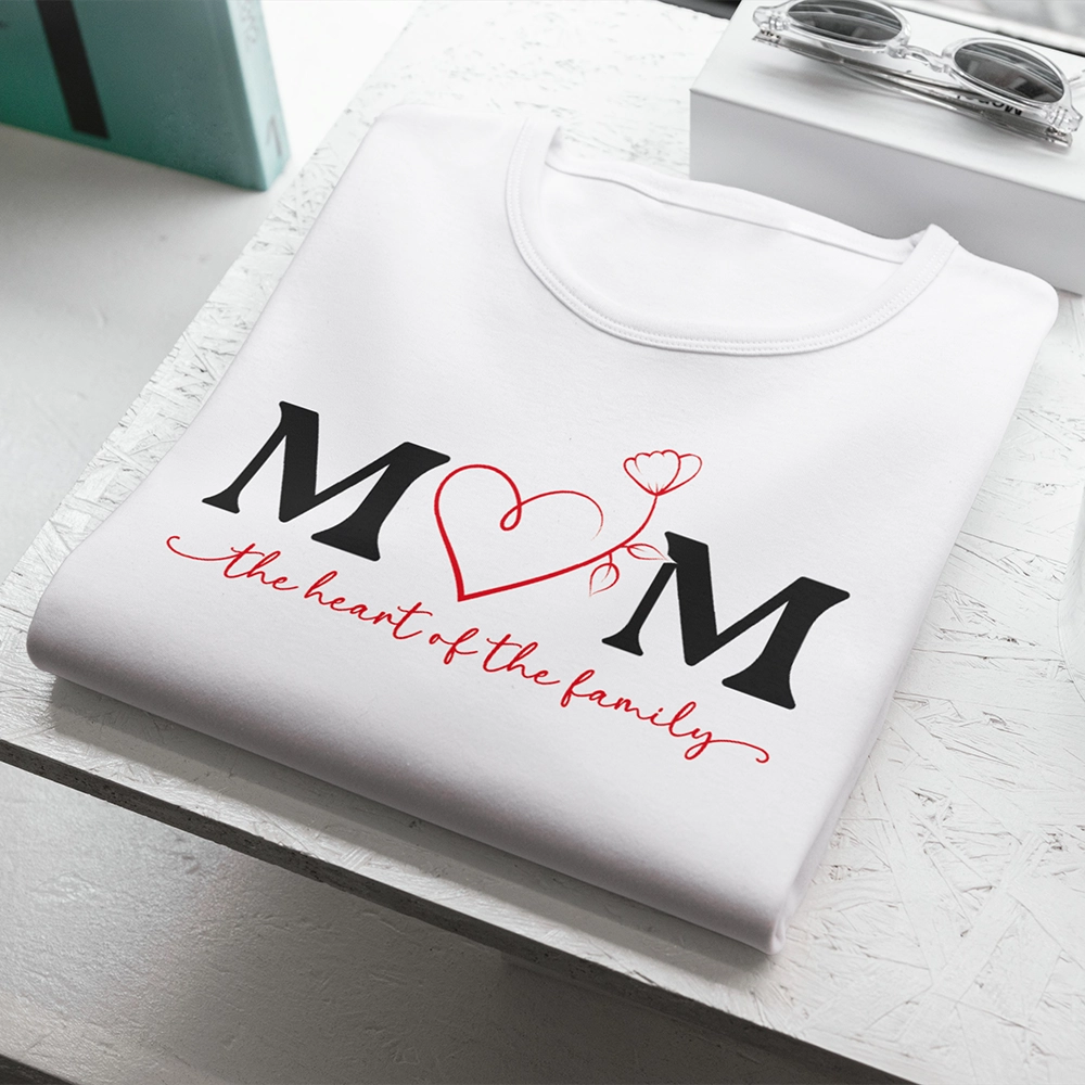 T-shirt bianca dedicata alla mamma con scritta Mom The Heart of the Family e cuore rosso centrale, piegata su piano chiaro.