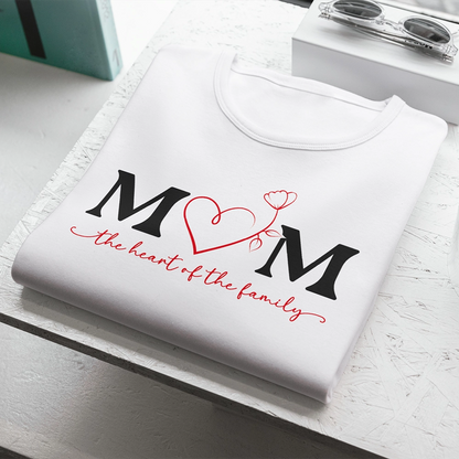 T-shirt bianca dedicata alla mamma con scritta Mom The Heart of the Family e cuore rosso centrale, piegata su piano chiaro.