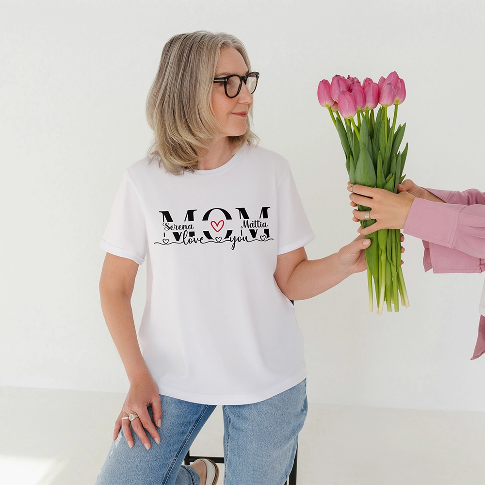 Donna con t-shirt bianca personalizzata per la mamma con nomi e dedica, mentre riceve un mazzo di tulipani rosa.