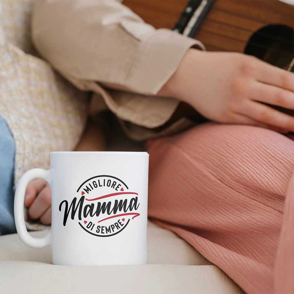 Tazza bianca dedicata alla mamma con scritta Migliore Mamma di sempre, ambientata su divano accanto a una persona seduta.