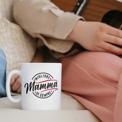 Tazza bianca dedicata alla mamma con scritta Migliore Mamma di sempre, ambientata su divano accanto a una persona seduta.