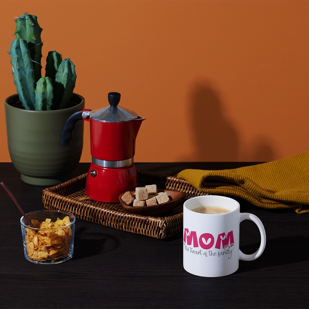 Tazza bianca con grafica Mom The Heart of the Family in ambientazione colazione con moka, cactus e cereali.