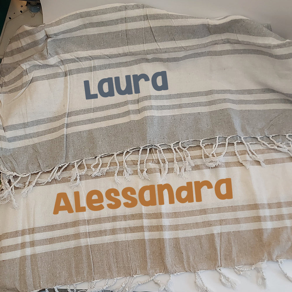 Teli mare fouta personalizzati nei colori grigio e tortora, con nomi stampati in contrasto e frange bianche.