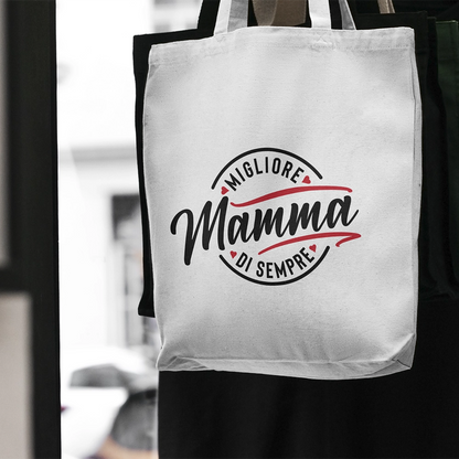 Tote bag bianca dedicata alla mamma con scritta Migliore Mamma di sempre, appesa in ambientazione minimal.