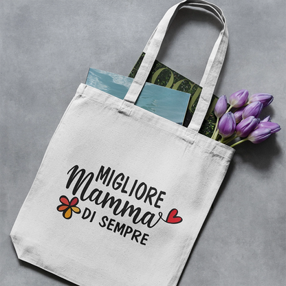 Tote bag bianca dedicata alla mamma con scritta Migliore Mamma di sempre, appoggiata con riviste e tulipani viola.