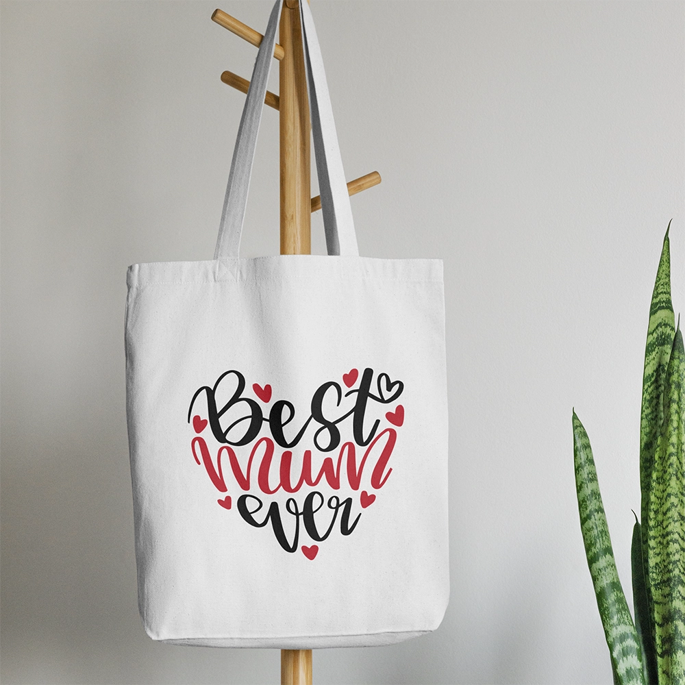 Tote bag bianca per la mamma con scritta Best Mum Ever e piccoli cuori rossi, appesa su supporto in legno.