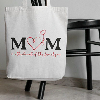 Tote bag bianca dedicata alla mamma con scritta Mom The Heart of the Family, appoggiata a una sedia in ambientazione indoor.